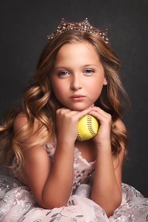 Young girl in a pink dress and jeweled crown holds a yellow softball, posing for a portrait against a dark background.
