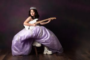 Young woman in lavender ball gown and tiara plays electric guitar while wearing white sneakers in studio portrait.