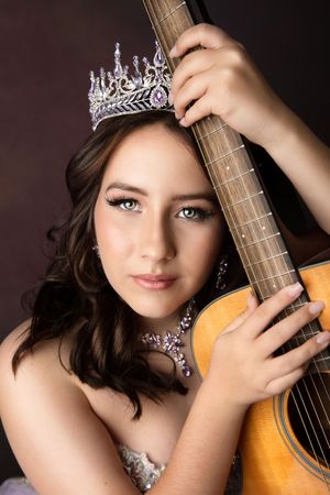 Young woman wearing a crystal tiara and jewelry poses elegantly with an acoustic guitar in a studio portrait.