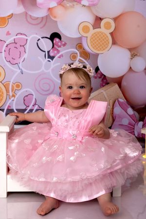 Baby in pink tulle dress poses against pastel balloon backdrop with Minnie Mouse decorations.