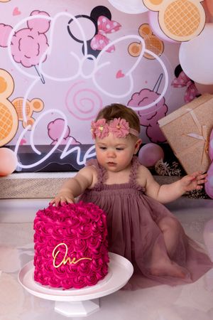 A smash cake photo session features a dark brown dress and bright pink cake on a white cake stand against floral backdrop.