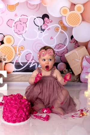 Baby wearing brown dress next to bright pink cake with decorative balloon backdrop in celebration setting.