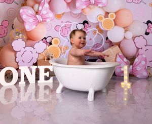 Baby's first birthday celebration setup with bathtub, cake, and pink balloon arch background.