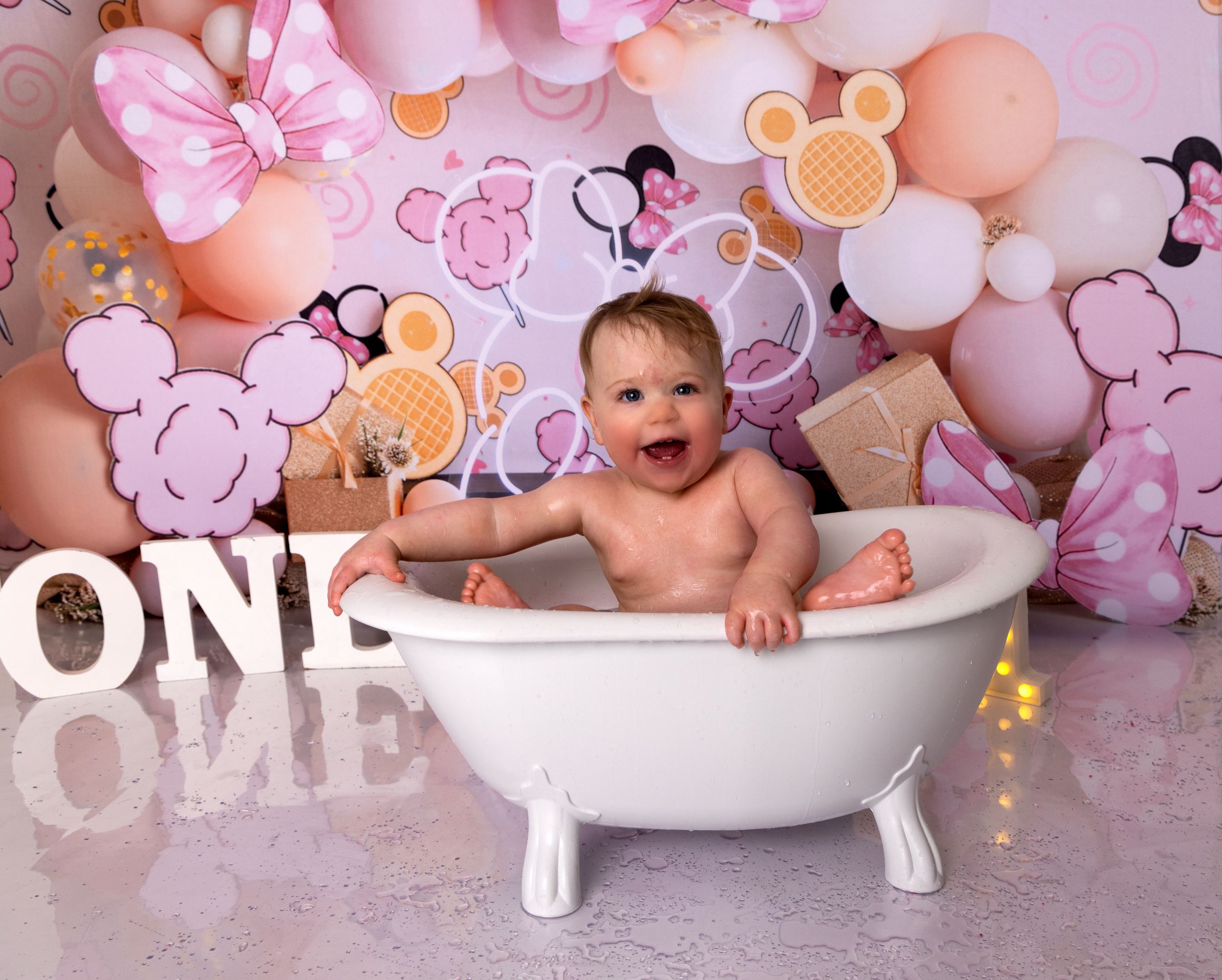 Baby bathtub photo shoot setup with pink and peach balloons, oranges and floral decorations against pink backdrop.