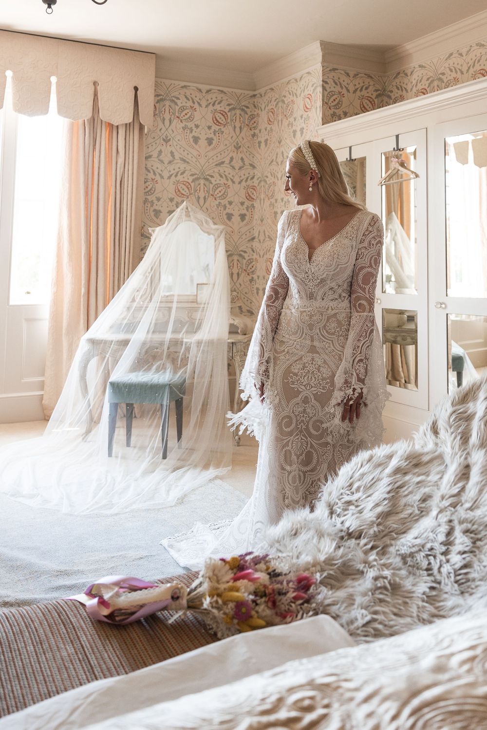 A lace wedding dress with long sleeves and intricate detailing shown in an elegant bedroom with floral wallpaper.