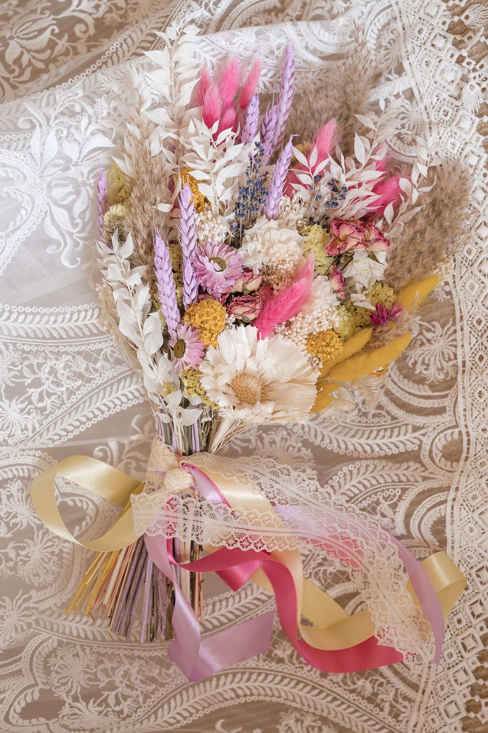 Delicate dried flower bouquet with pink and cream blooms arranged with ribbons against a textured lace background.