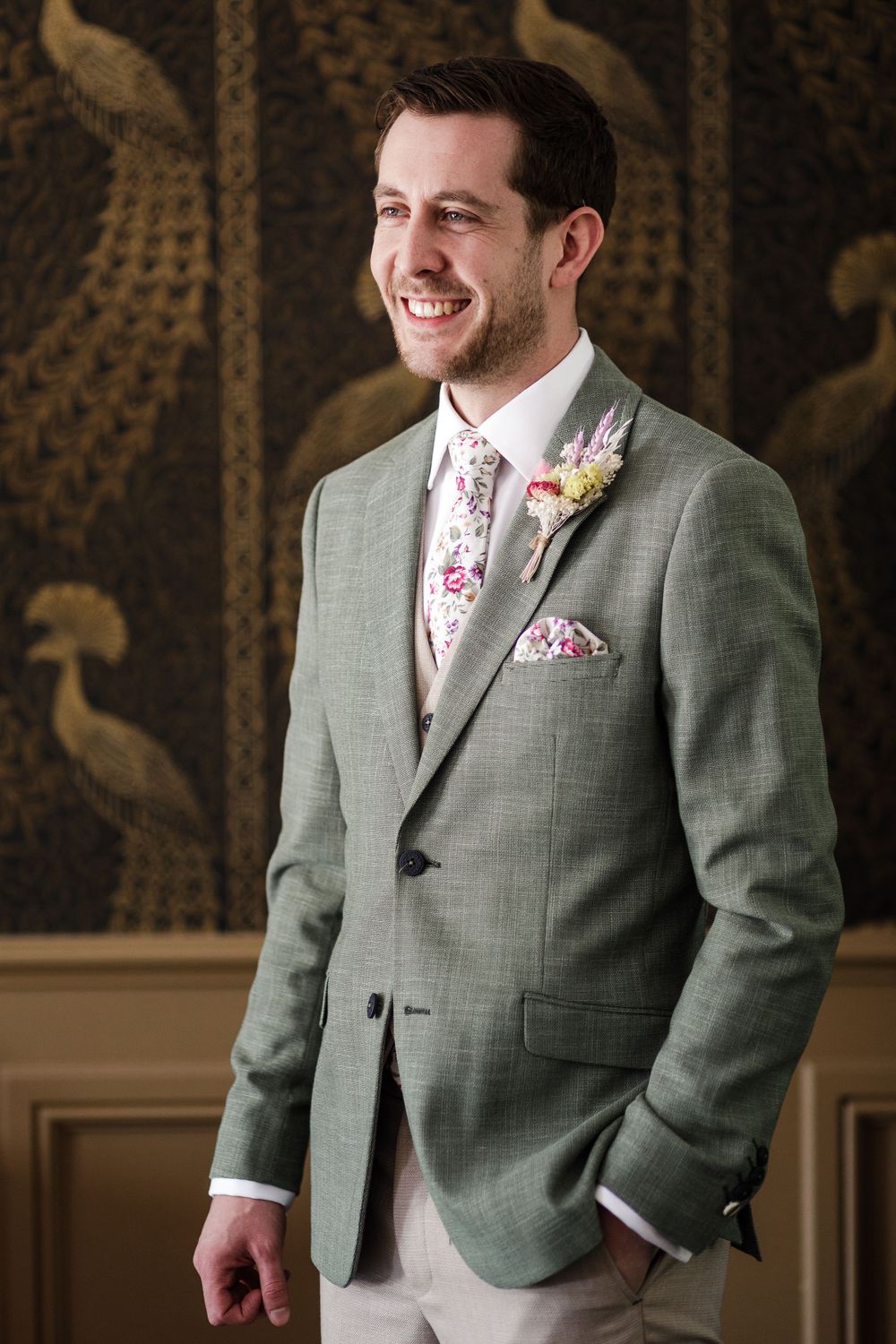 Person wearing light gray suit jacket with floral tie and pocket square smiles warmly against dark patterned wallpaper.