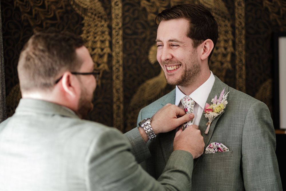 Wedding party members in tailored gray suits laugh together against an ornate dark backdrop.