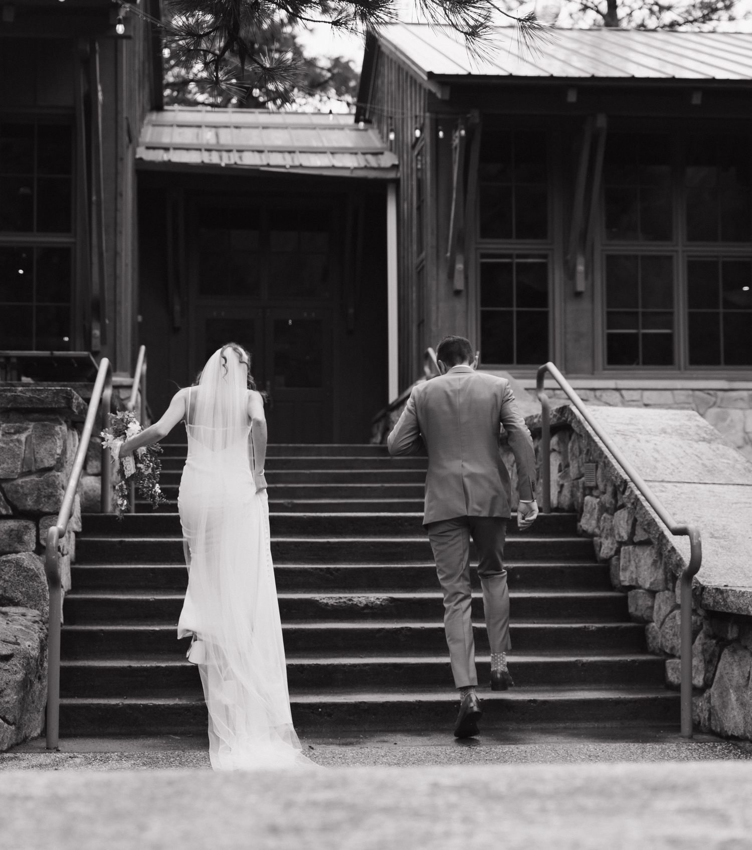 A black and white photo of newlyweds ascending stone stairs to a rustic lodge entrance.