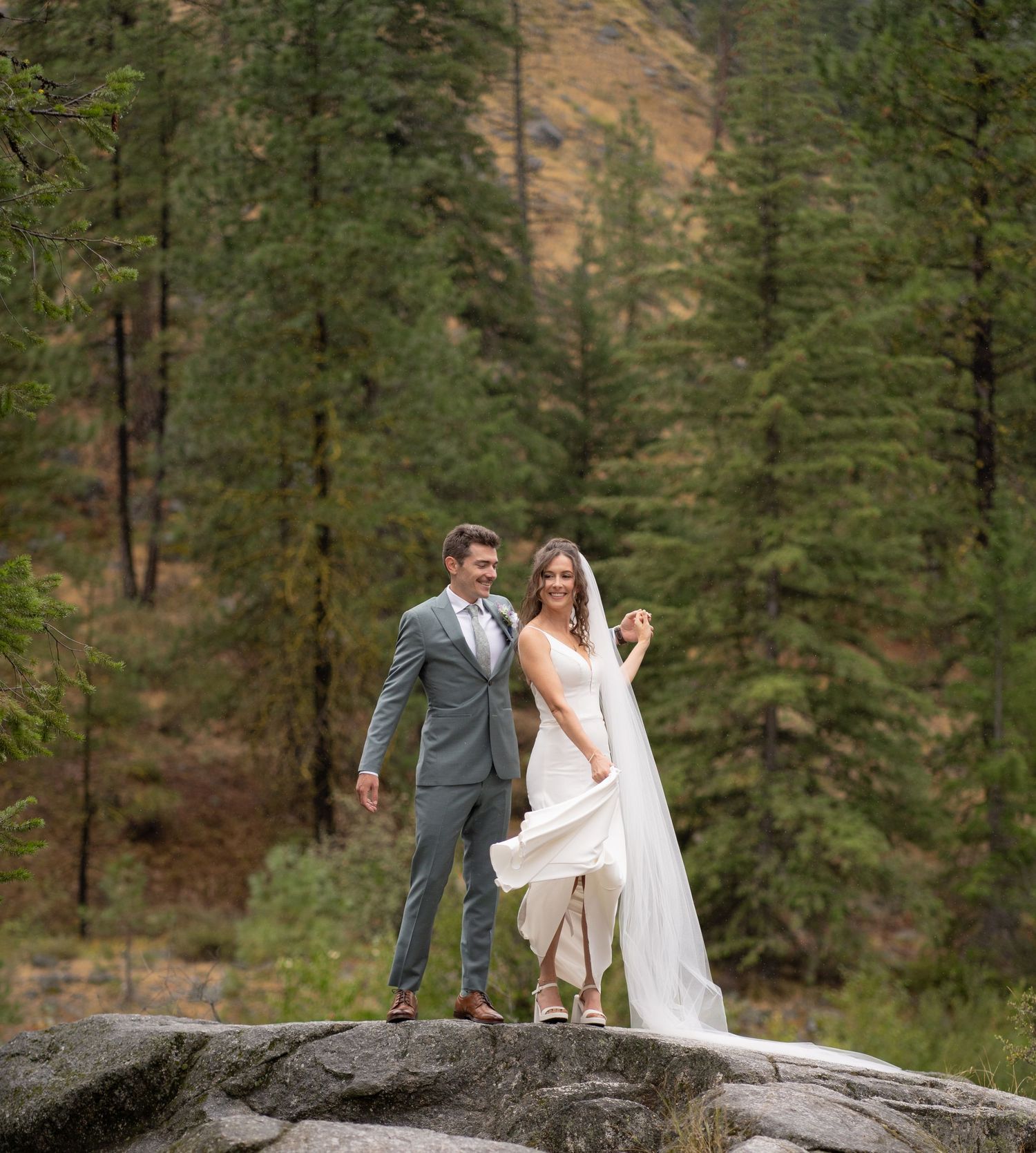 Newlyweds share a joyful moment while standing on a rocky outcrop surrounded by pine forest.