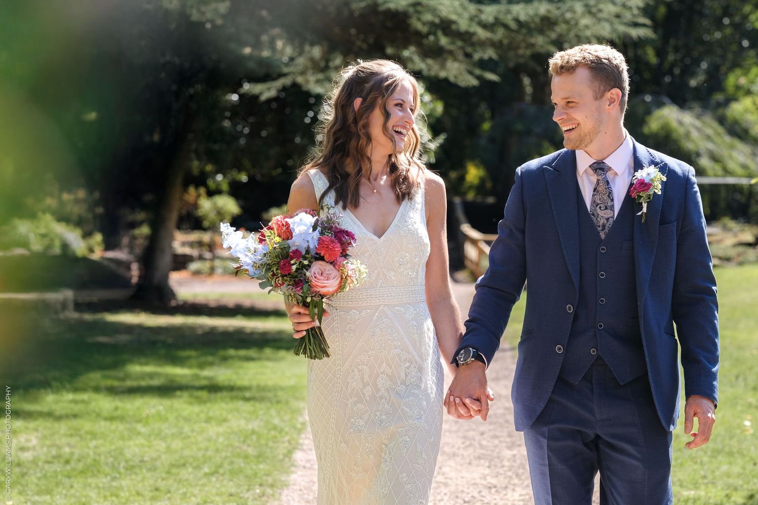 A couple walks hand in hand across a lush garden lawn in elegant wedding attire.