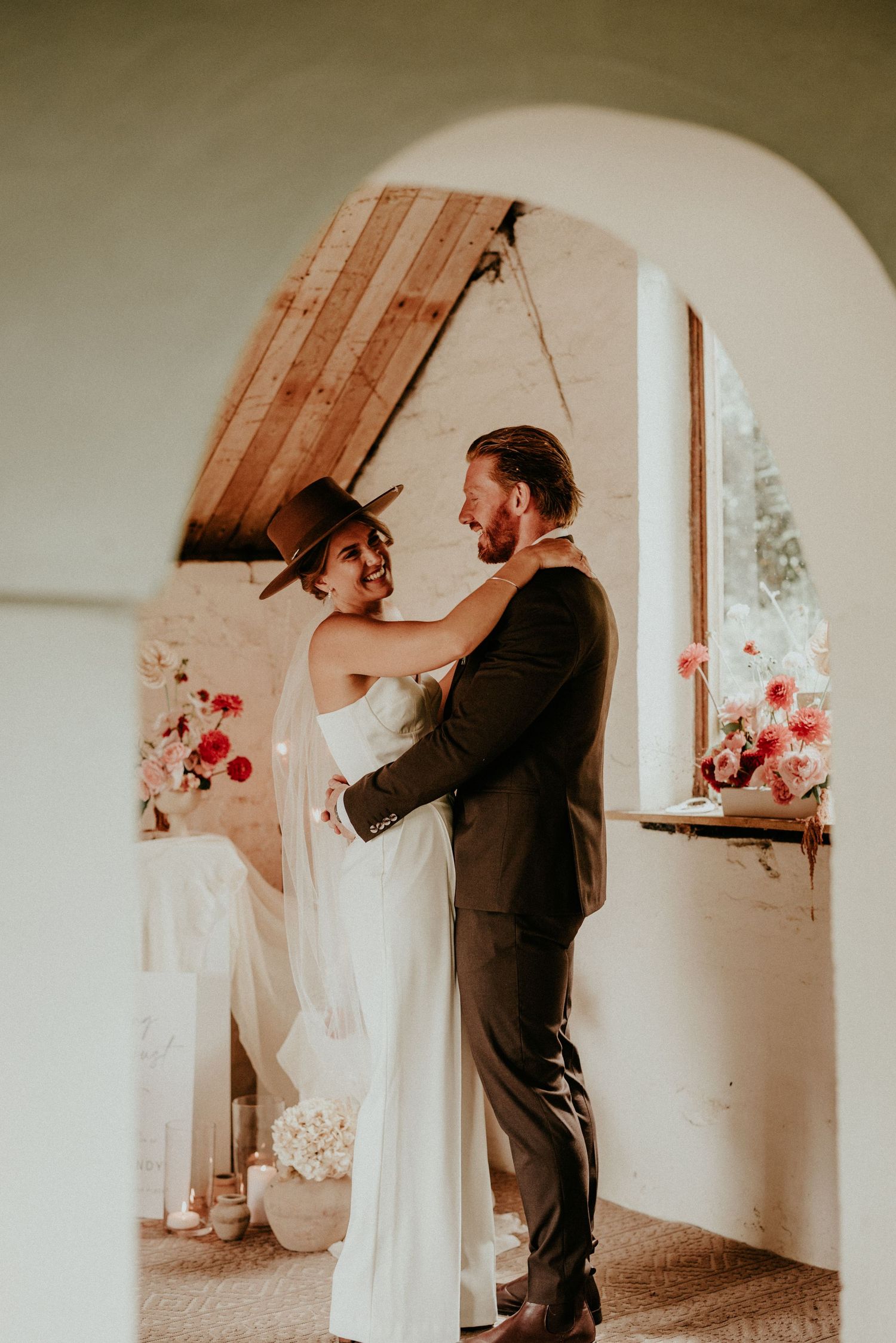 A romantic first look moment shared by a couple in a white-walled room with natural window light and red floral accents.