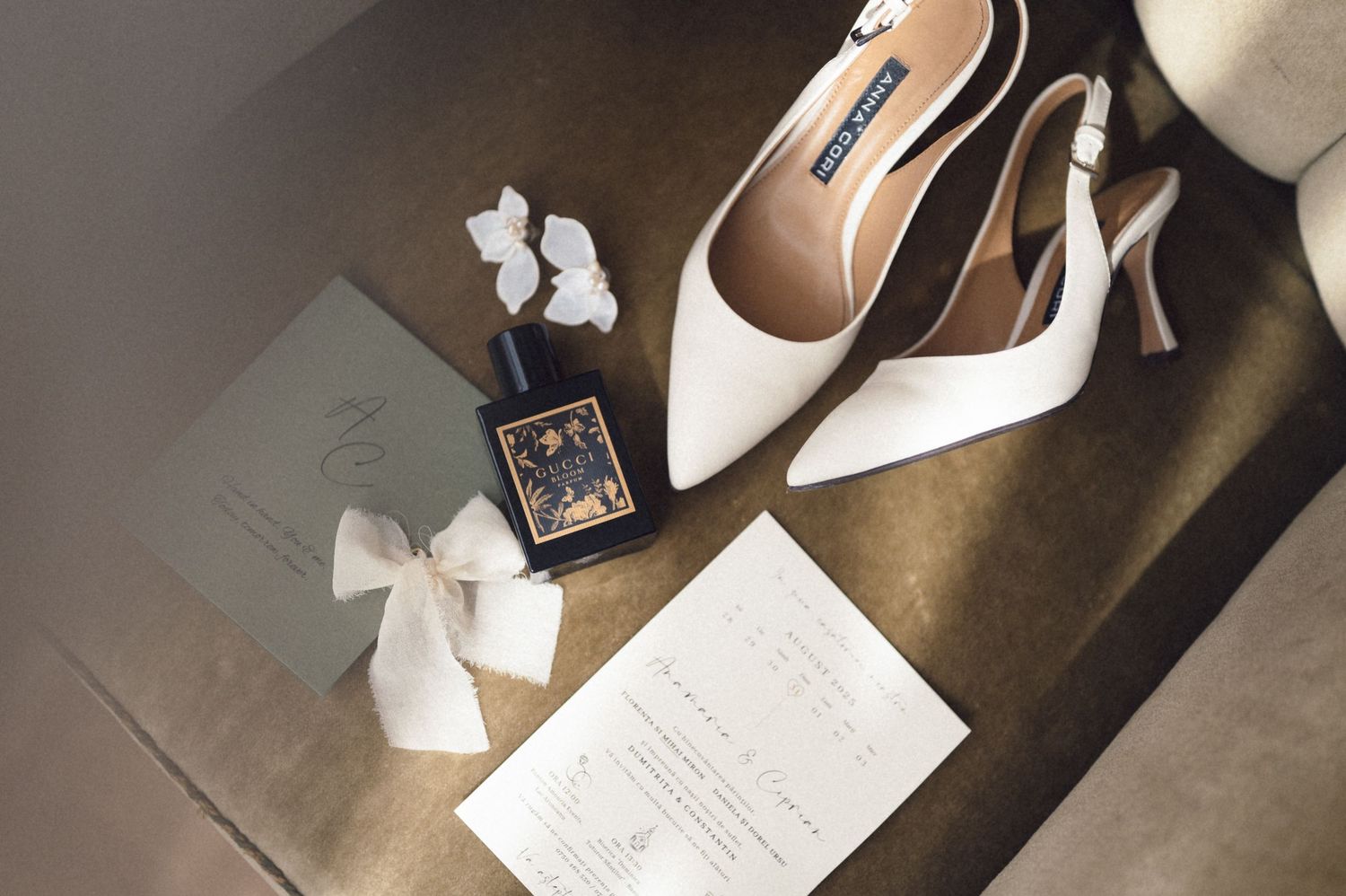 Wedding accessories including white slingback heels, perfume bottle, invitation and hair bow arranged on dark surface.