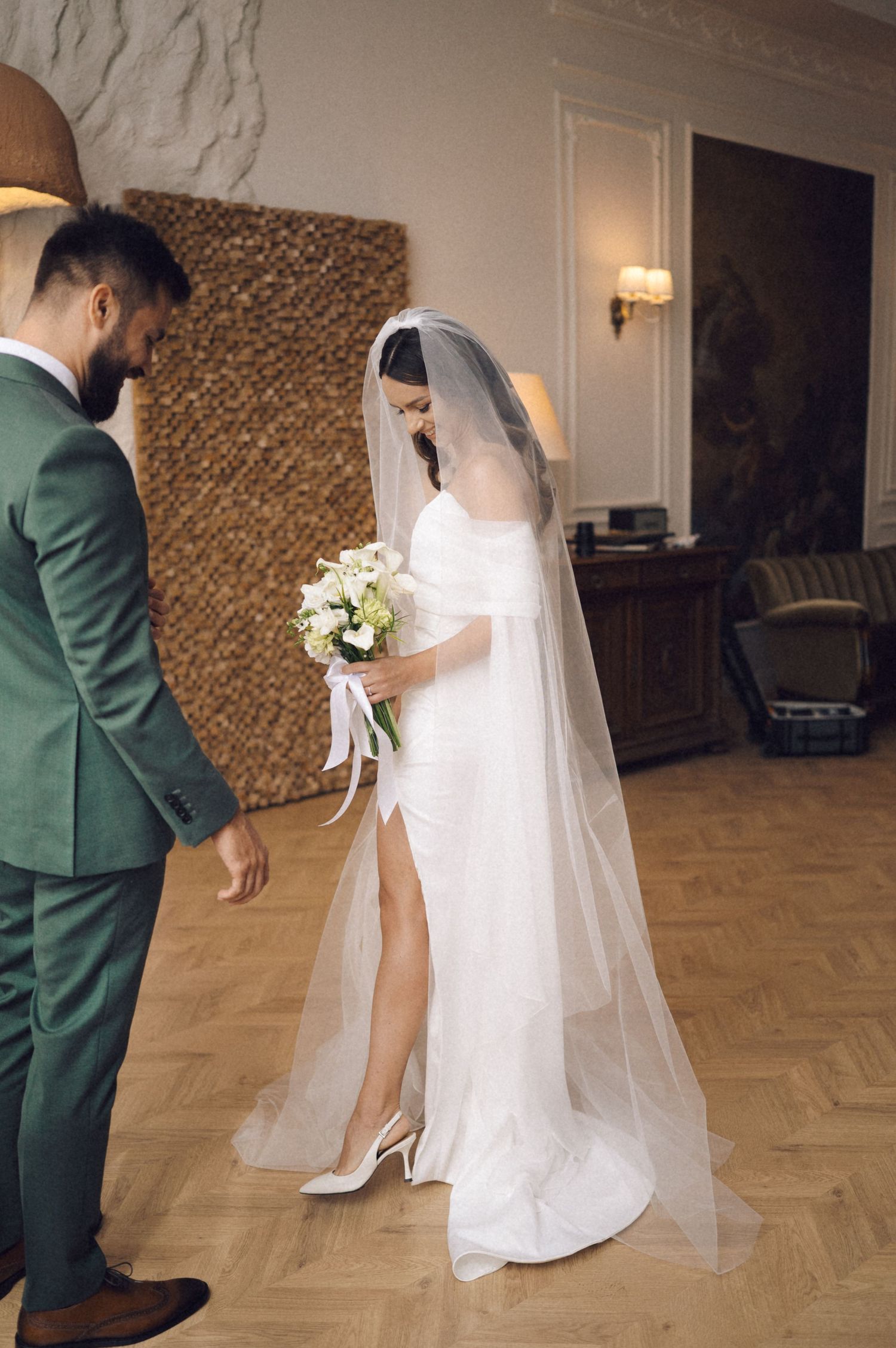 A bride in white dress and veil dances with groom in green suit on a parquet dance floor.