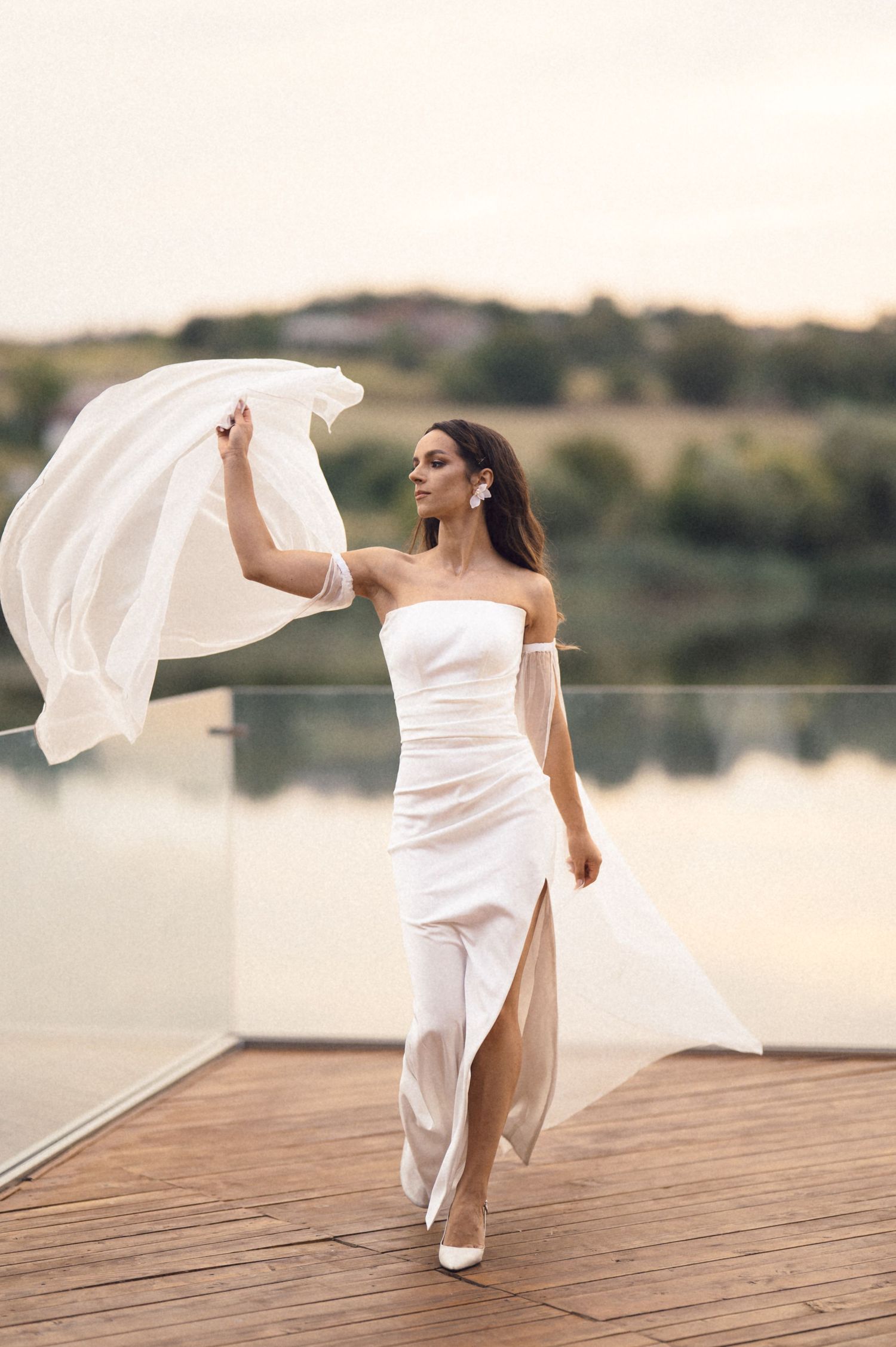 A woman in a flowing white dress dances gracefully on a wooden dock by a serene lake at sunset.