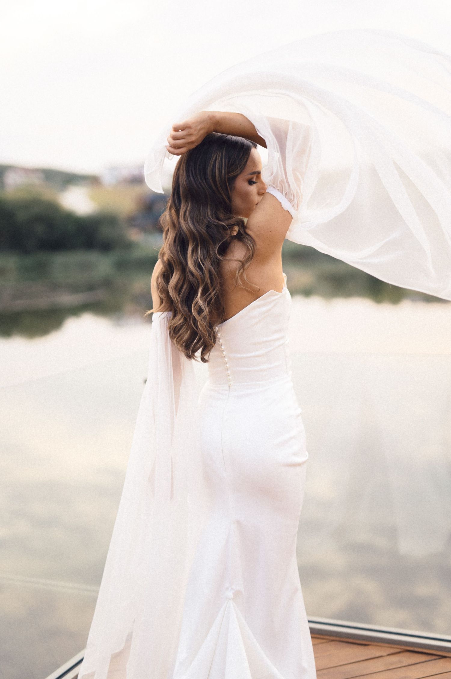 A bride in a flowing white dress and veil poses elegantly against a scenic outdoor backdrop.