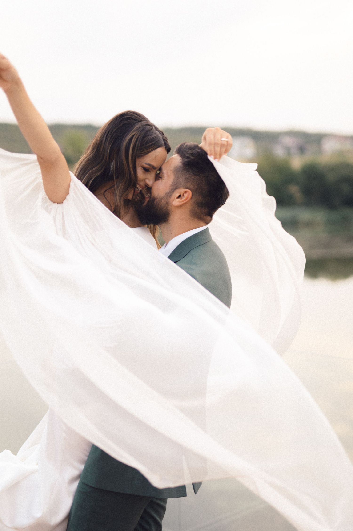 A romantic wedding couple embracing outdoors with flowing white veil caught in the breeze.