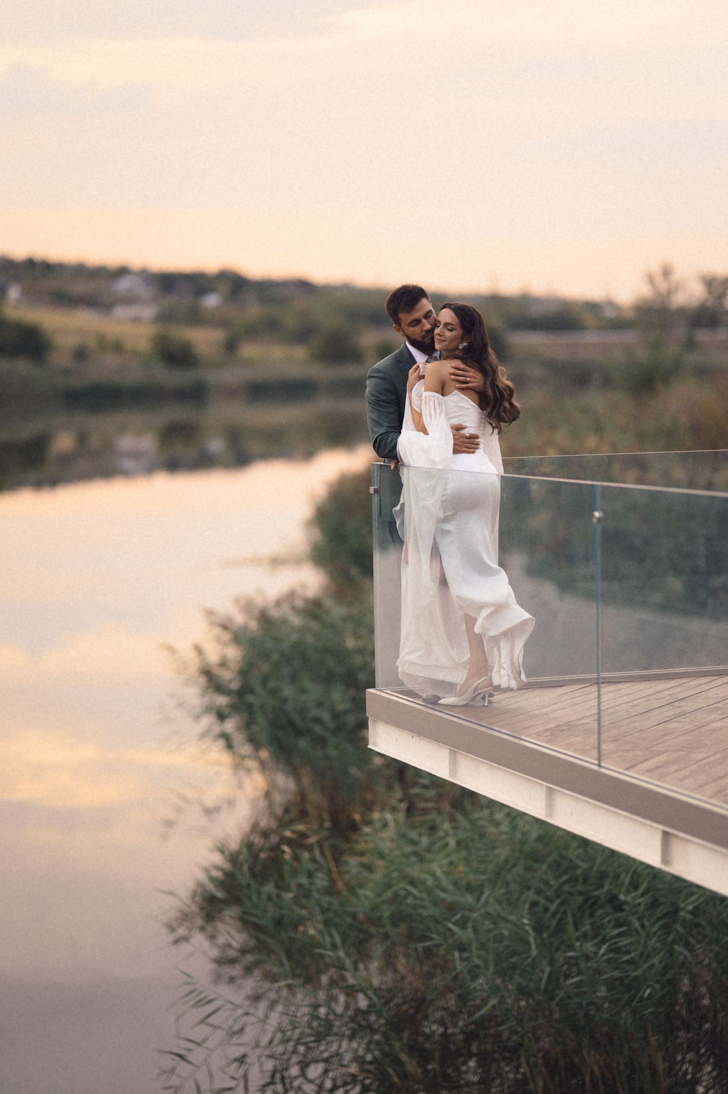 A couple in white outfits embraces on a wooden deck overlooking a serene lake during sunset.