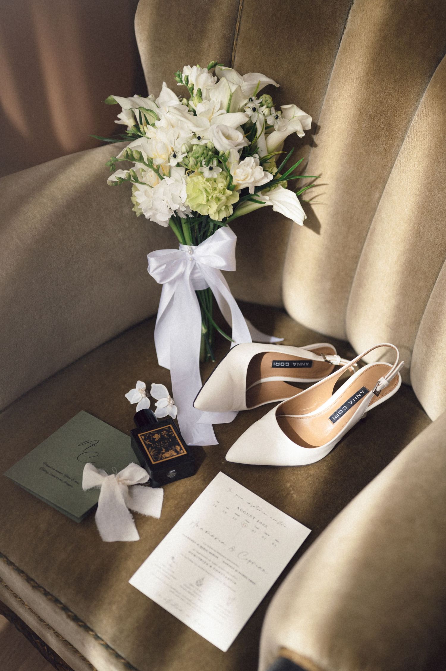 White floral bouquet, elegant high heel shoes, and handwritten note arranged on beige fabric in soft natural lighting.