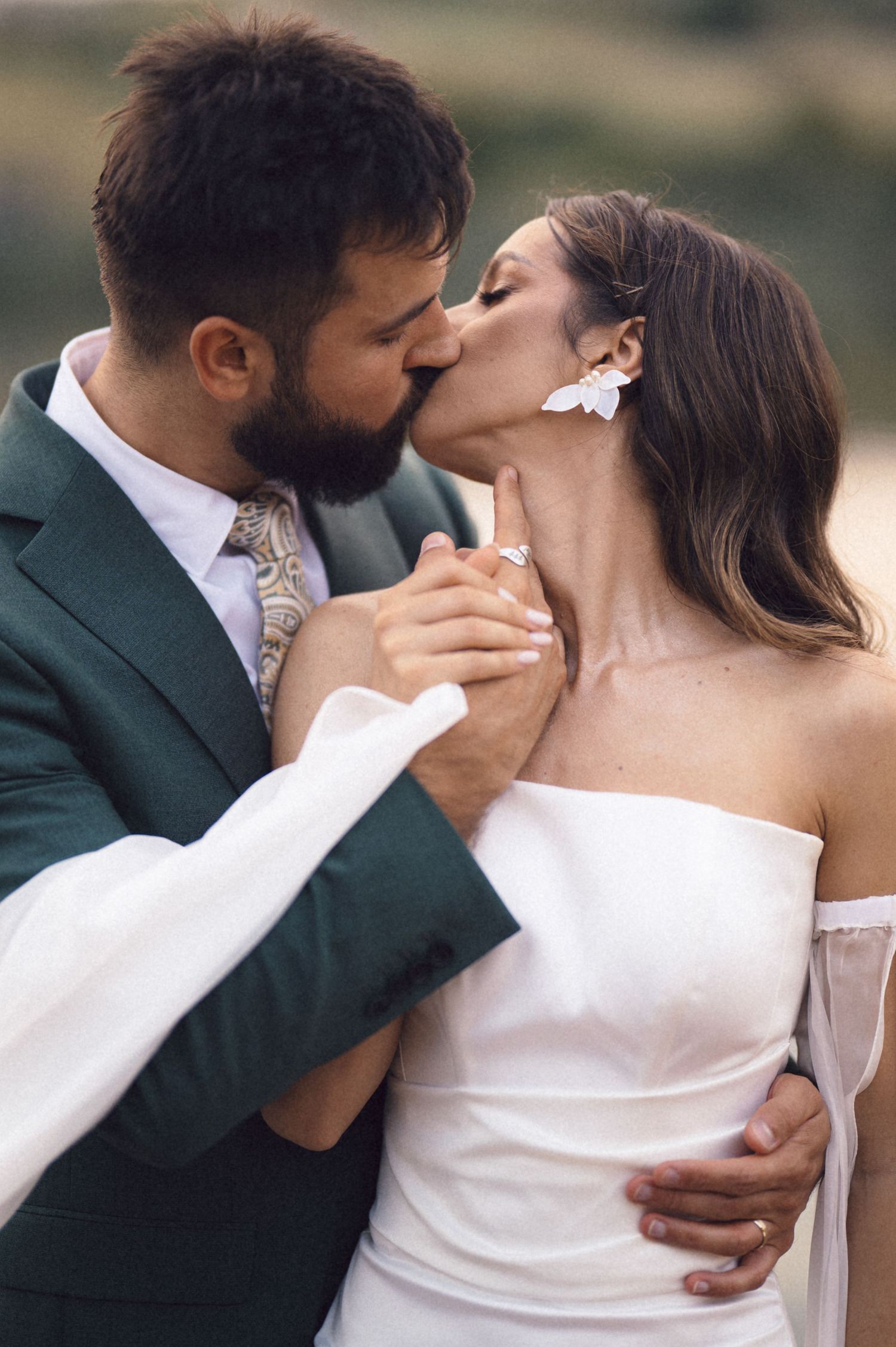 A romantic wedding couple sharing an intimate kiss while embracing outdoors, with the groom in a dark green suit.