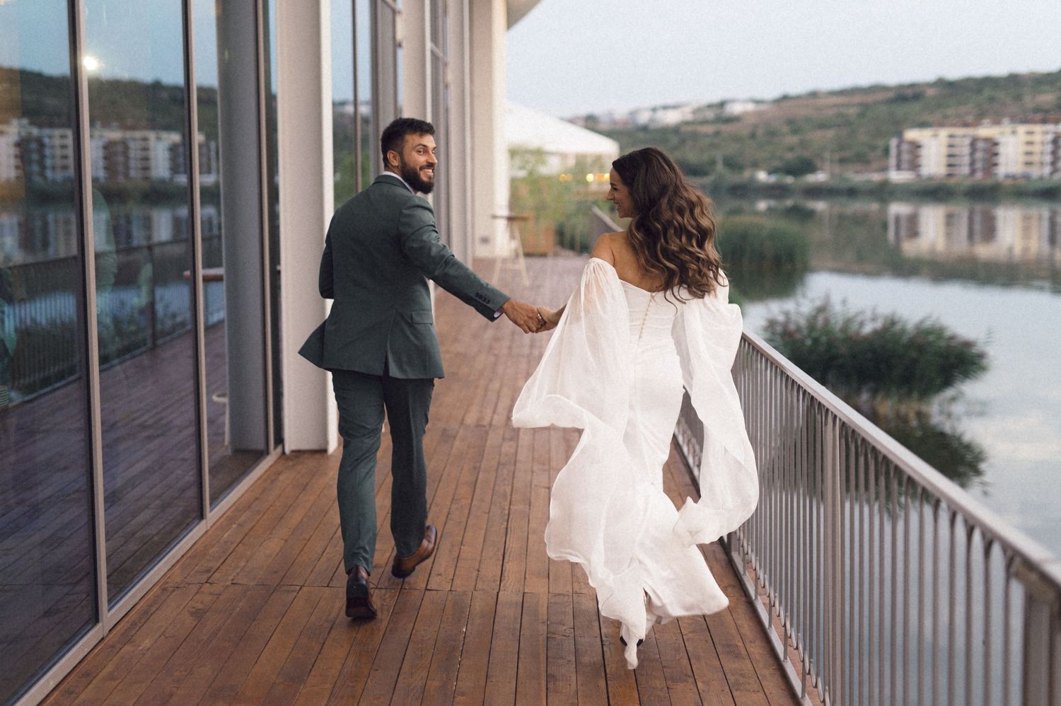 A couple walks and dances together on a wooden deck balcony overlooking a waterfront view.
