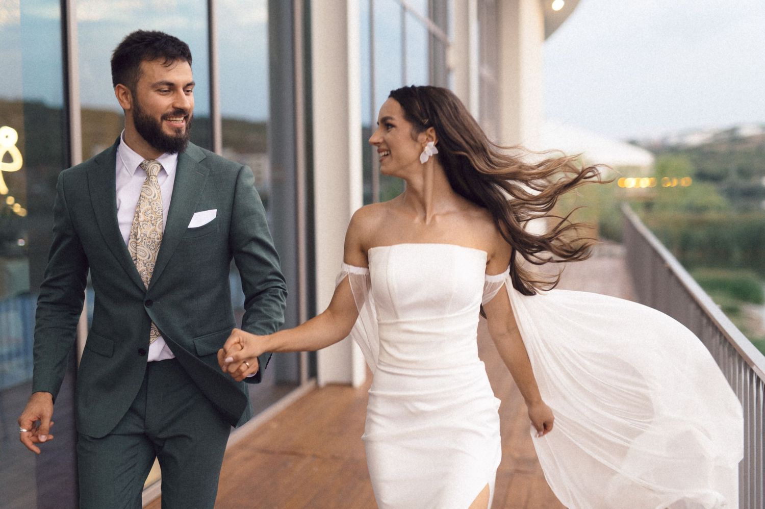 A couple in wedding attire walk together on a balcony, with the bride's white dress flowing in the breeze.