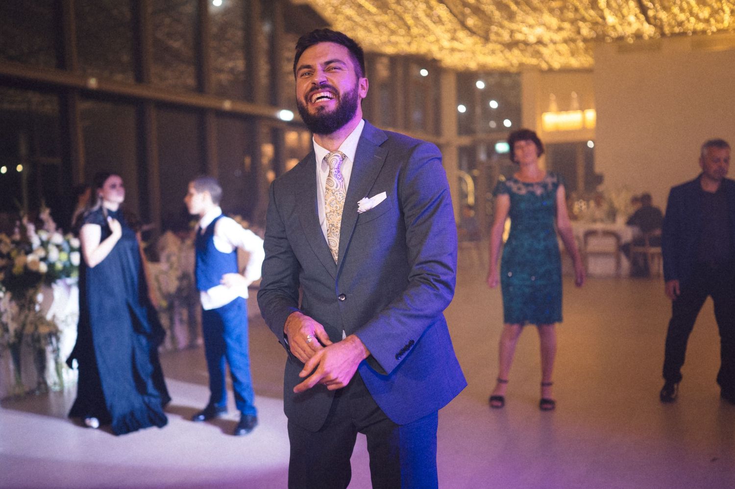 A group of wedding guests dance and celebrate on a dimly lit dance floor at an evening reception.