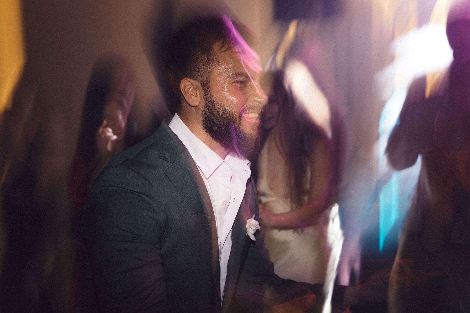 A group of people dancing and celebrating at a dimly lit party or nightclub with colorful lights in the background.