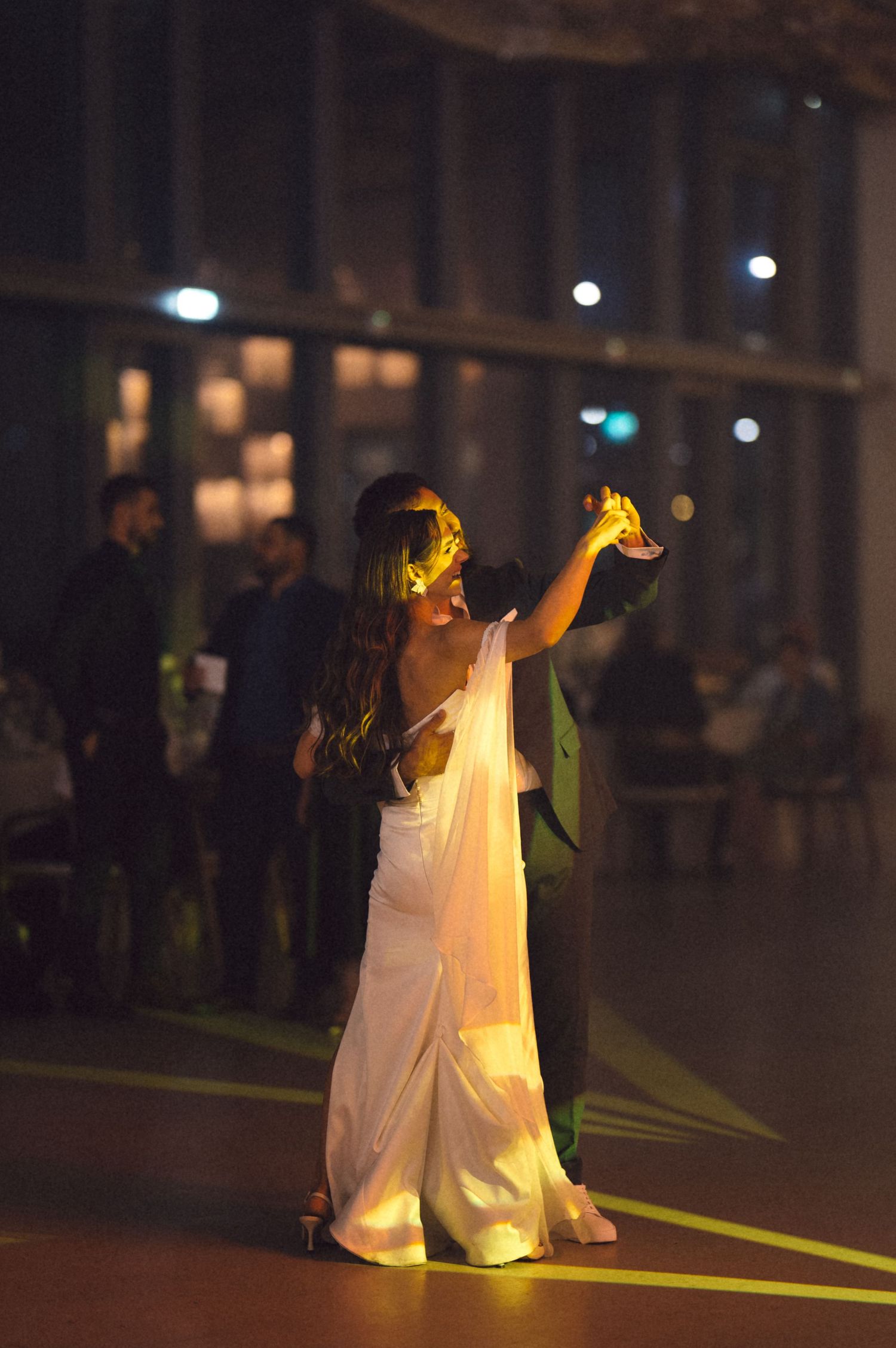 A couple dances together at a wedding reception, with the bride in a white gown illuminated in the dark venue.