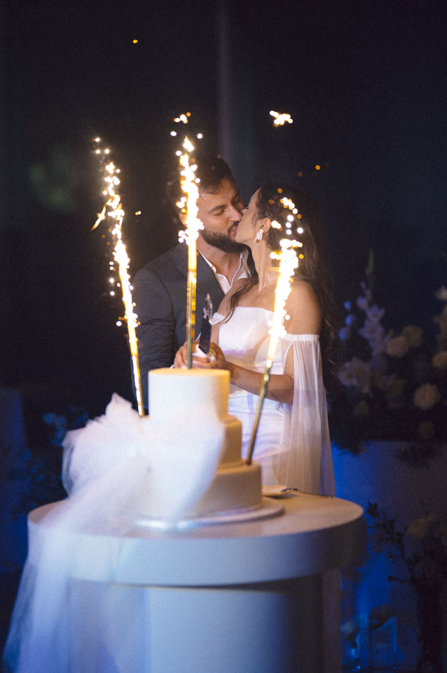 A wedding couple shares a romantic moment beside their cake with sparkler fountains illuminating the dark reception venue.