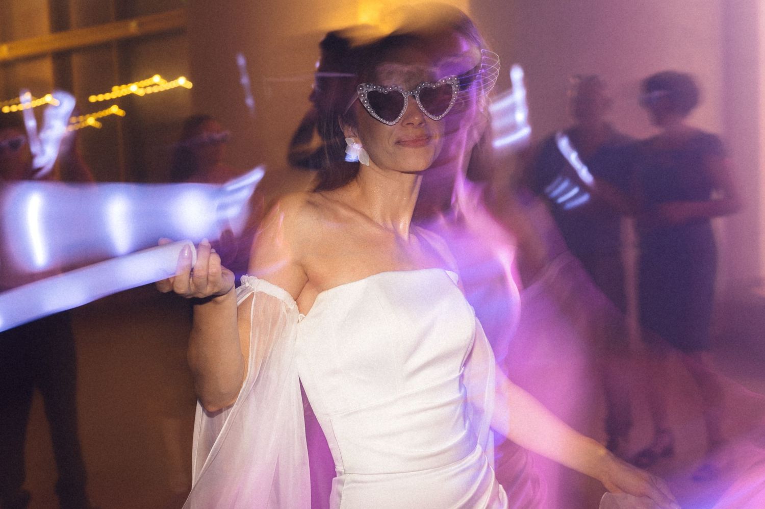 A group of wedding guests dancing and celebrating on a dimly lit dance floor with purple lighting effects.