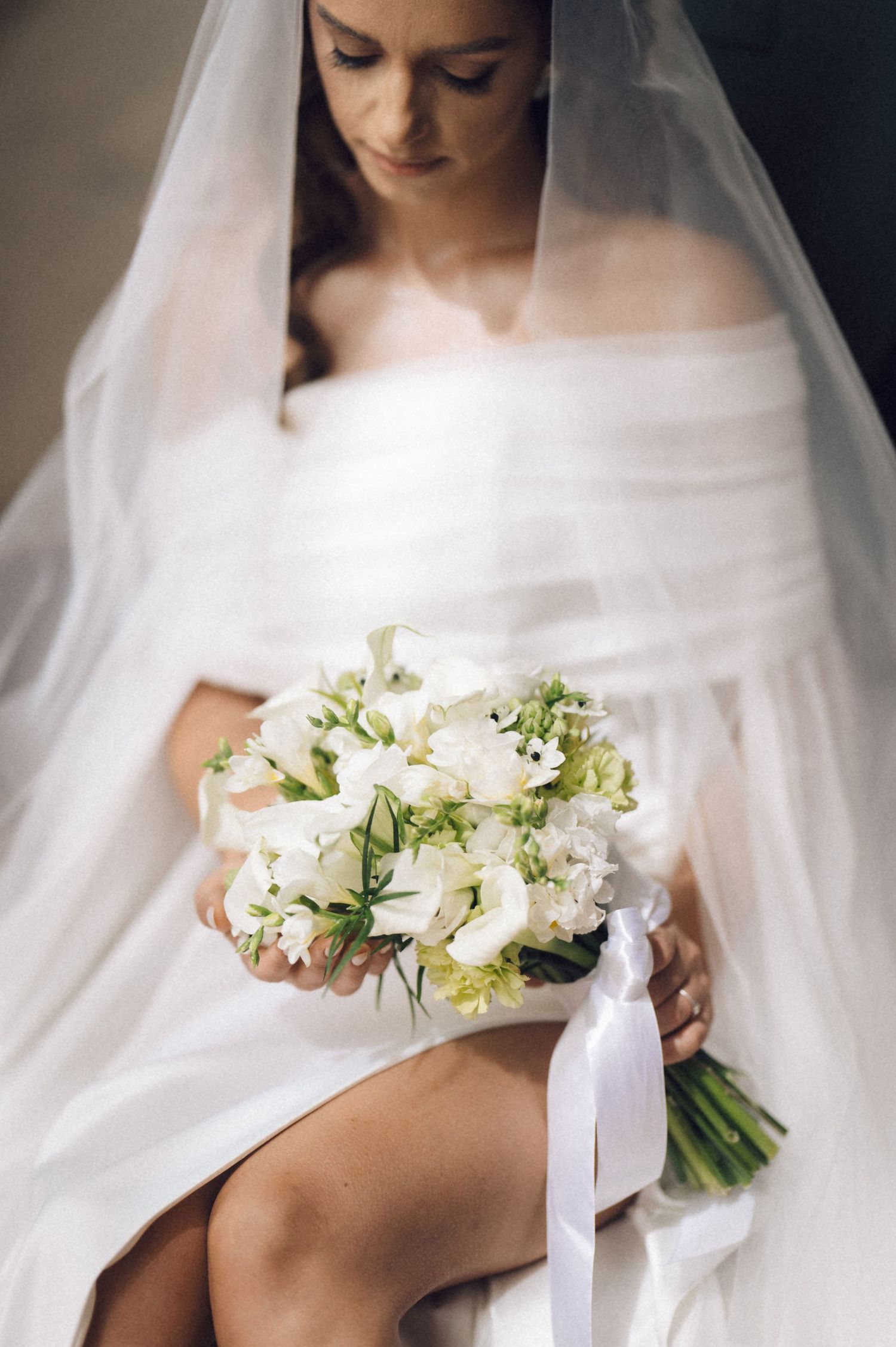 A bride in a white wedding dress holds an elegant bouquet of white flowers against a flowing veil background.