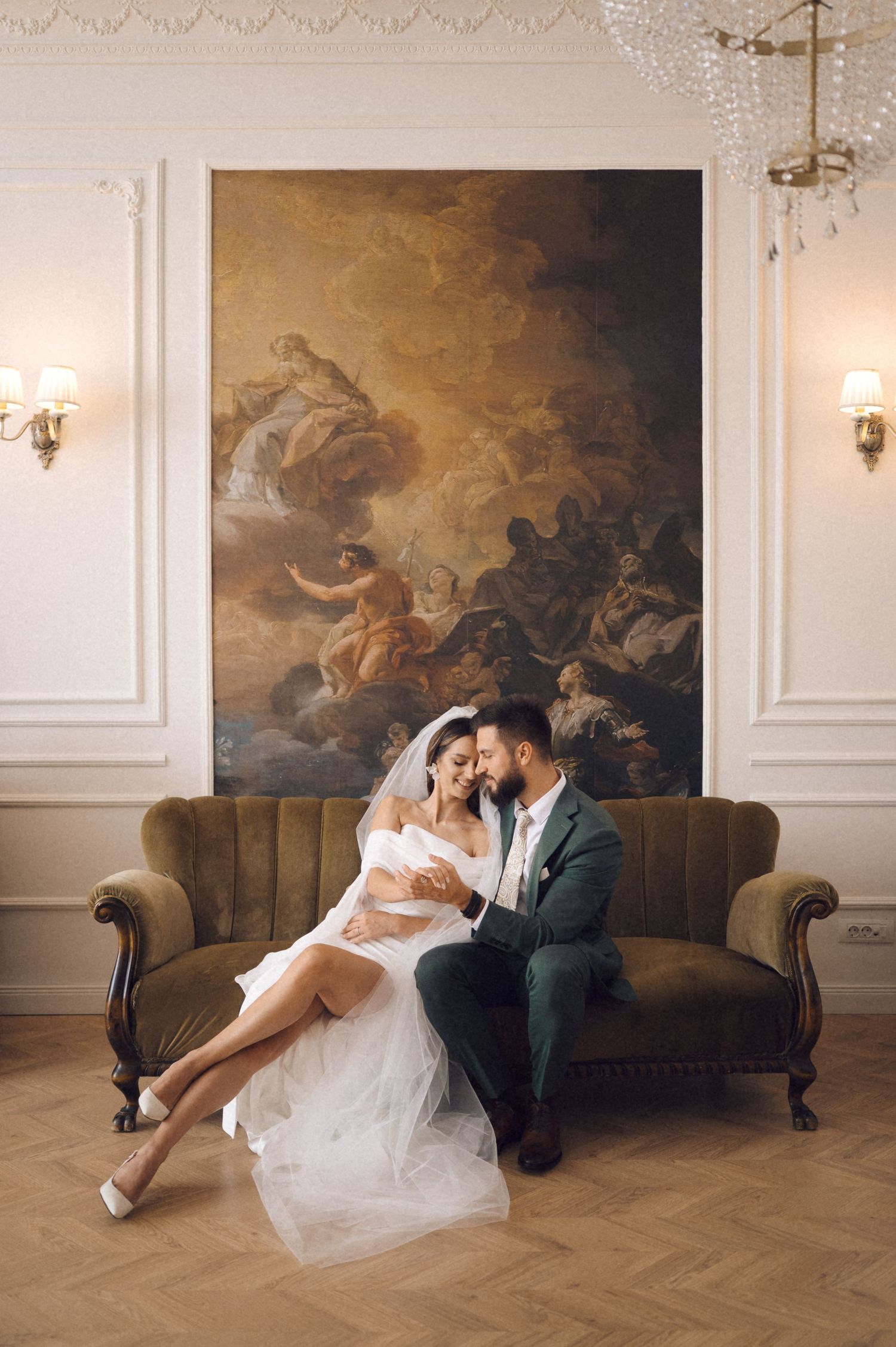 Elegant couple sits intimately on vintage brown sofa beneath ornate classical wall painting and sconces in refined interior.