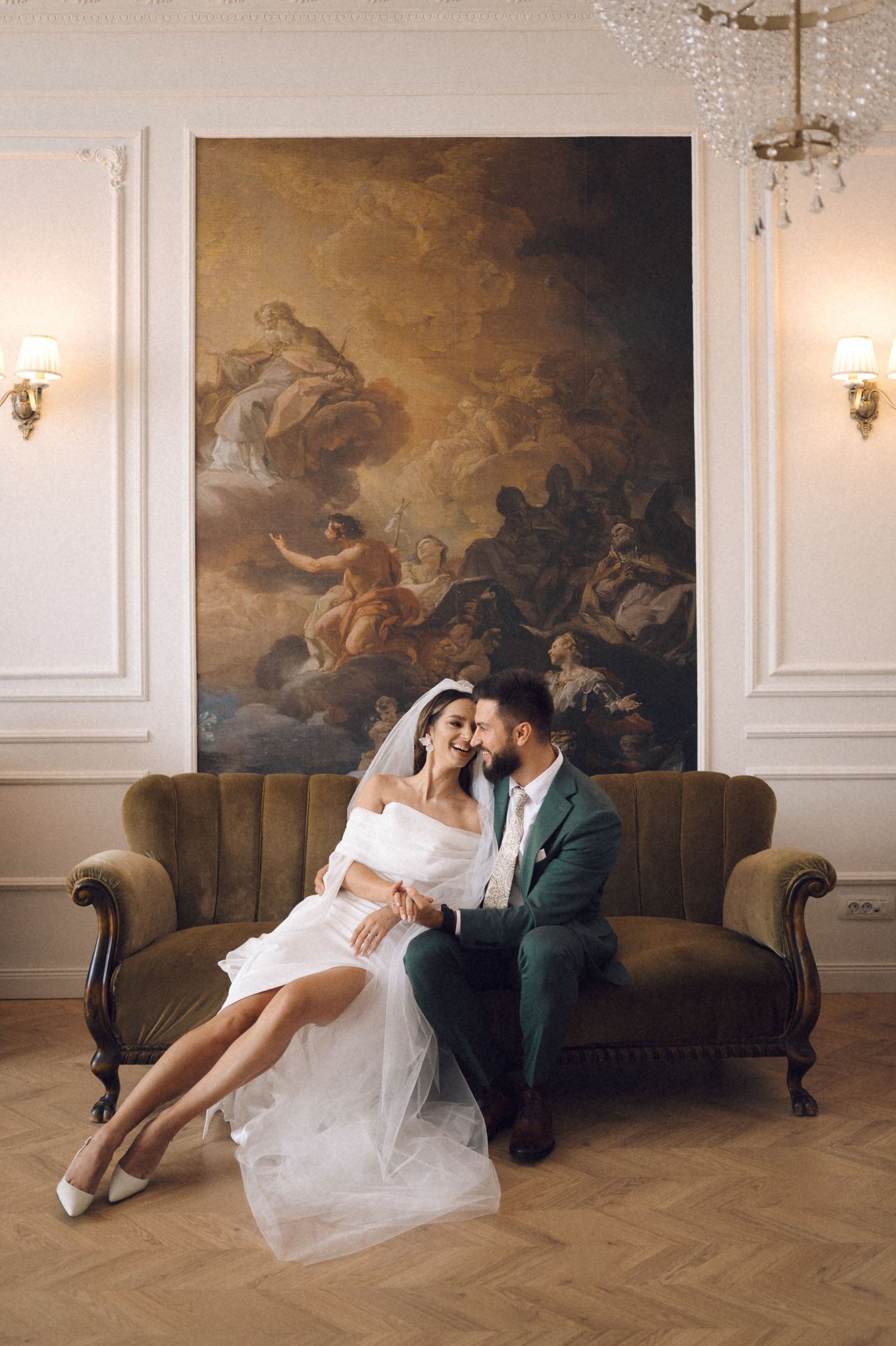 Romantic wedding couple lounges together on vintage sofa against ornate wall with classical painting and sconces.