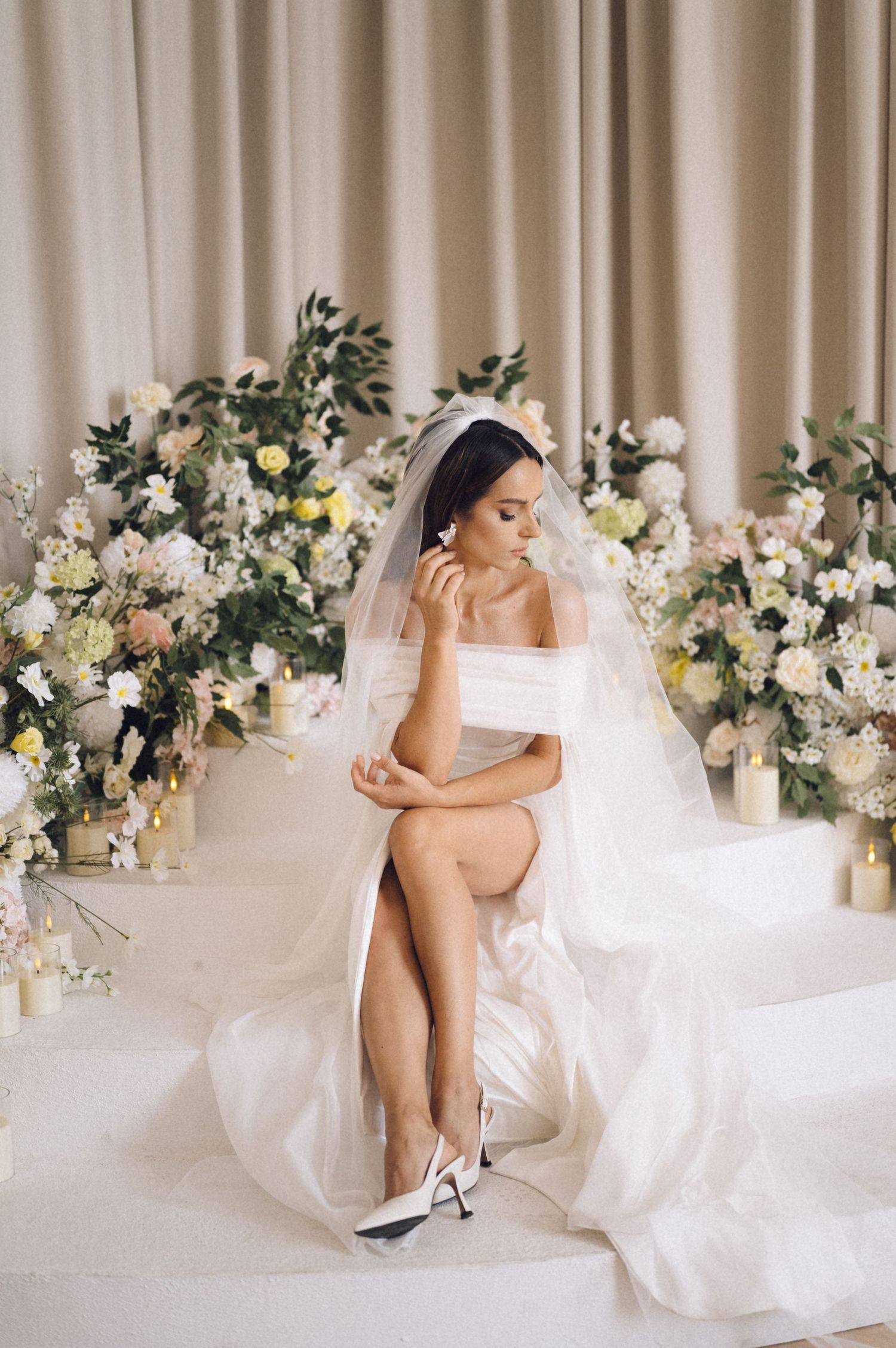 A bride in a flowing white wedding gown and heels sits elegantly in a room decorated with white and yellow floral arrangements.