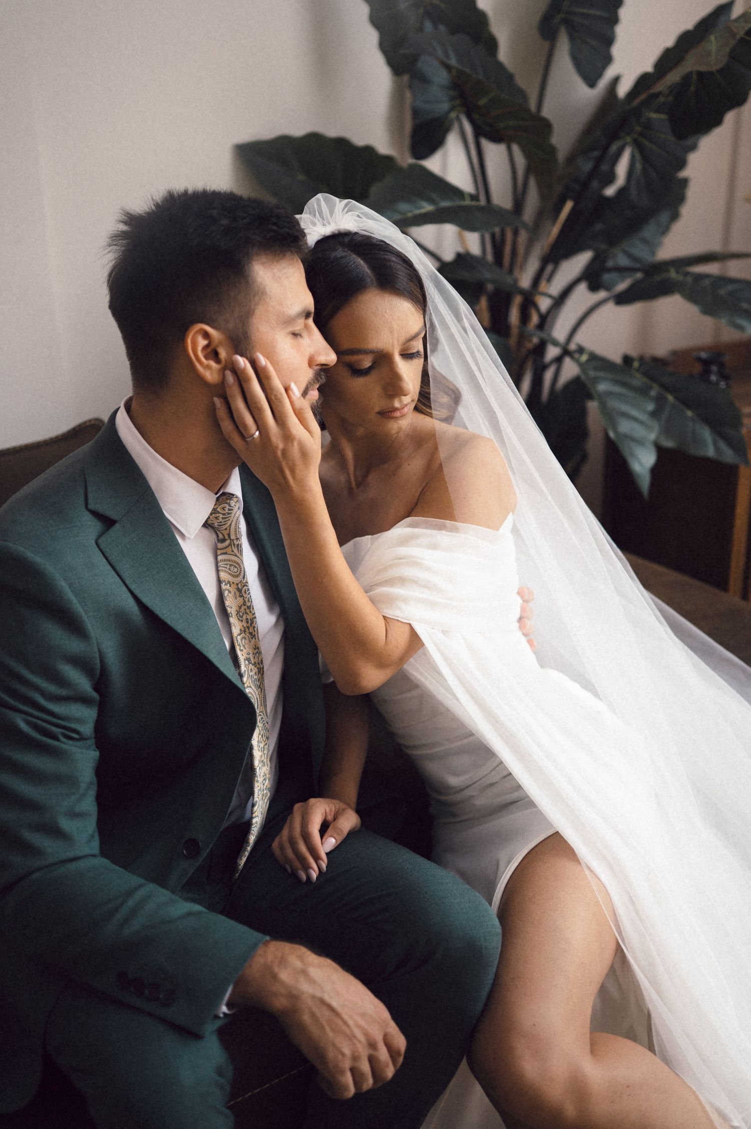 A couple in wedding attire share an intimate moment on a couch with a green plant in the background.