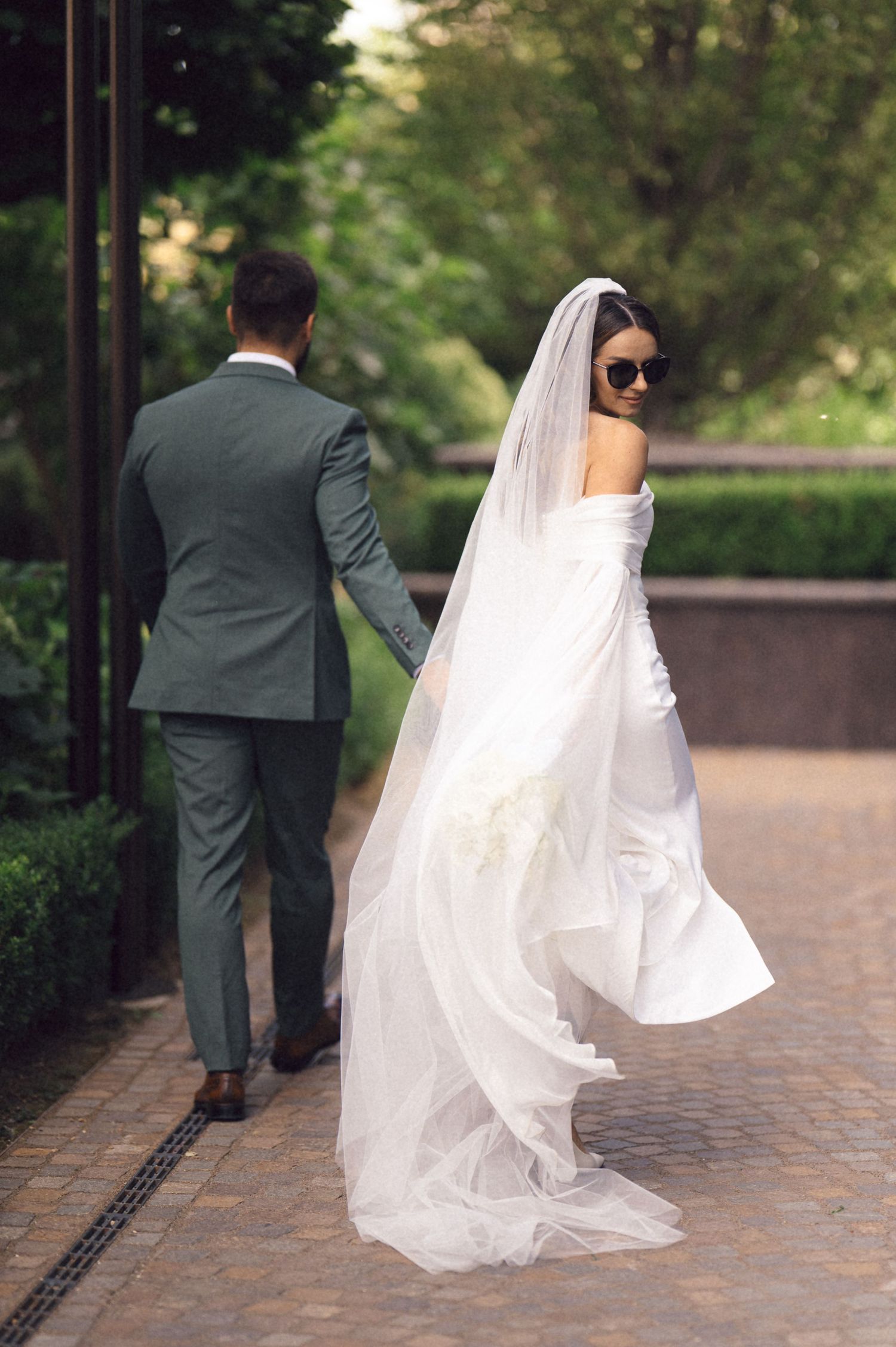 A bride and groom walk along a brick pathway with flowing white veil trailing behind them in an outdoor garden setting.