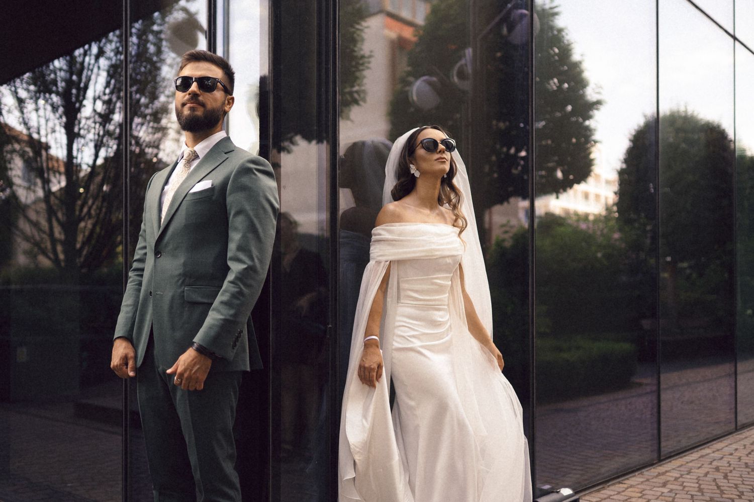 A couple walks together outdoors, with one wearing a green suit and the other in an elegant white dress.
