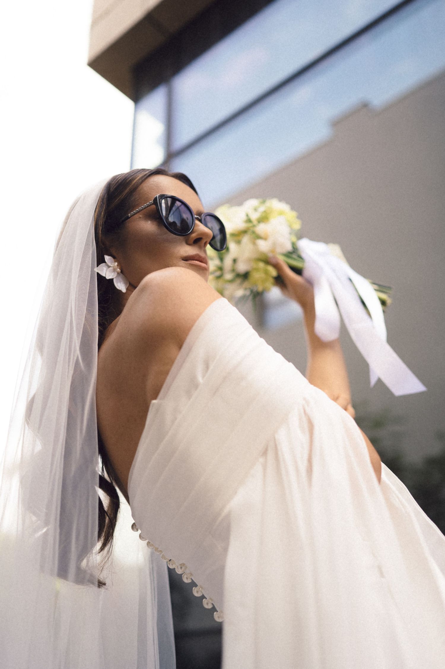 A bride in a white wedding dress and veil poses dramatically against a bright background during a photoshoot.