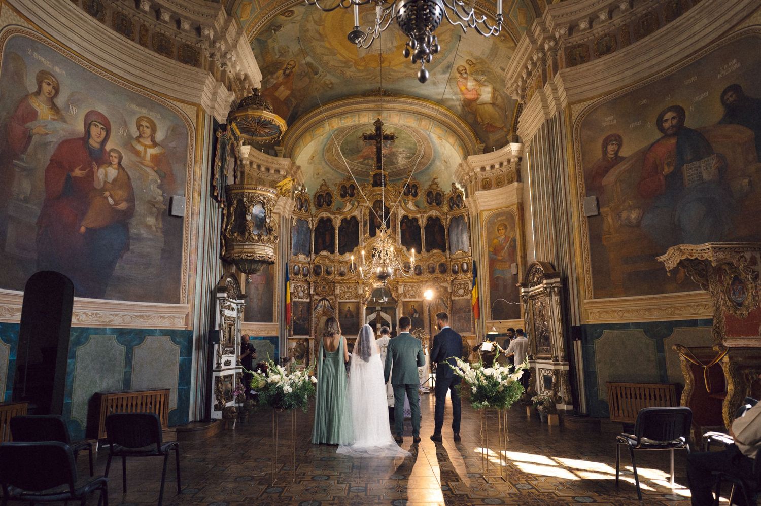 Ornate church interior with crystal chandeliers, religious artwork, and a wedding ceremony in progress.