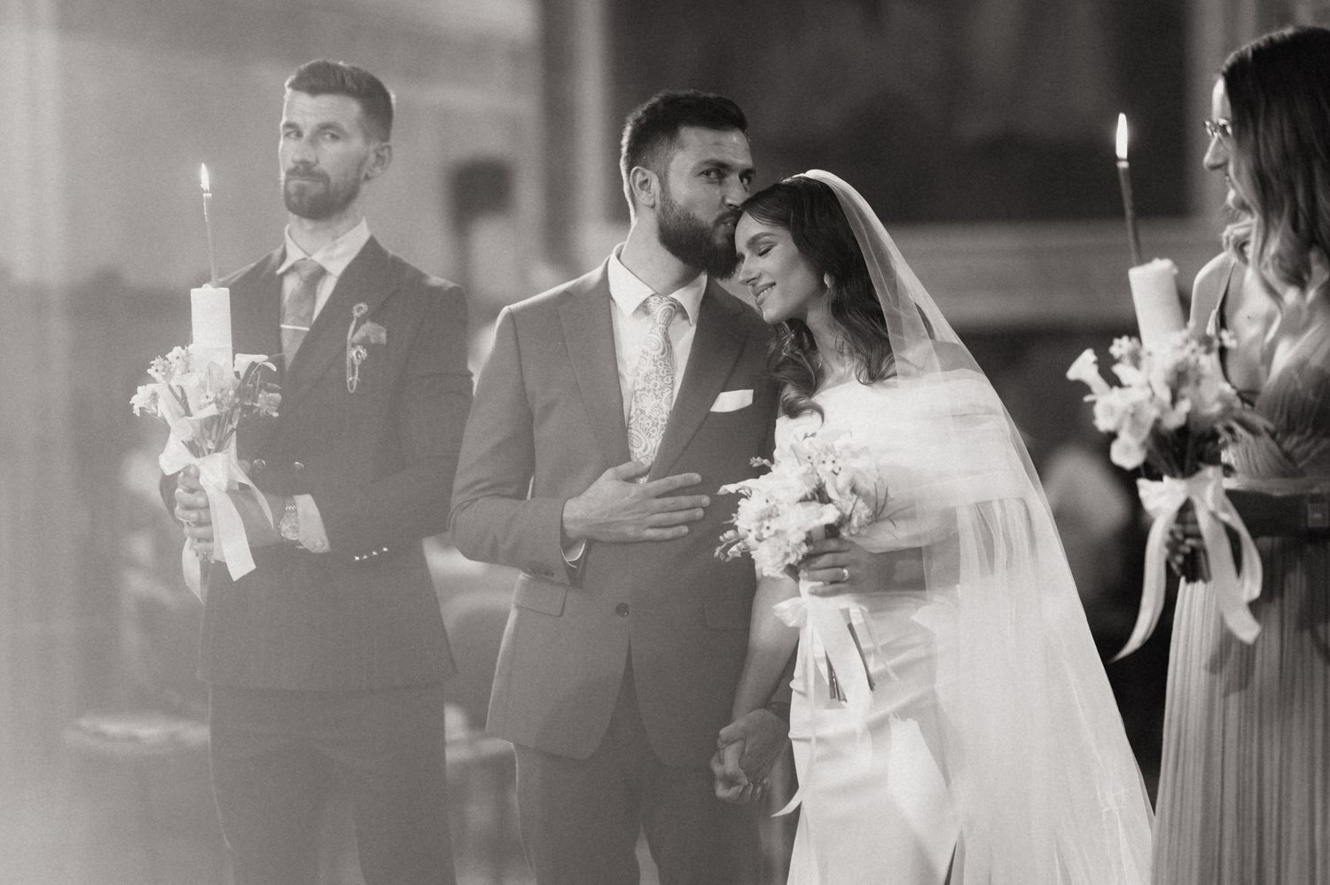Black and white wedding ceremony photograph showing a bride and groom with bridesmaids holding candles in church.