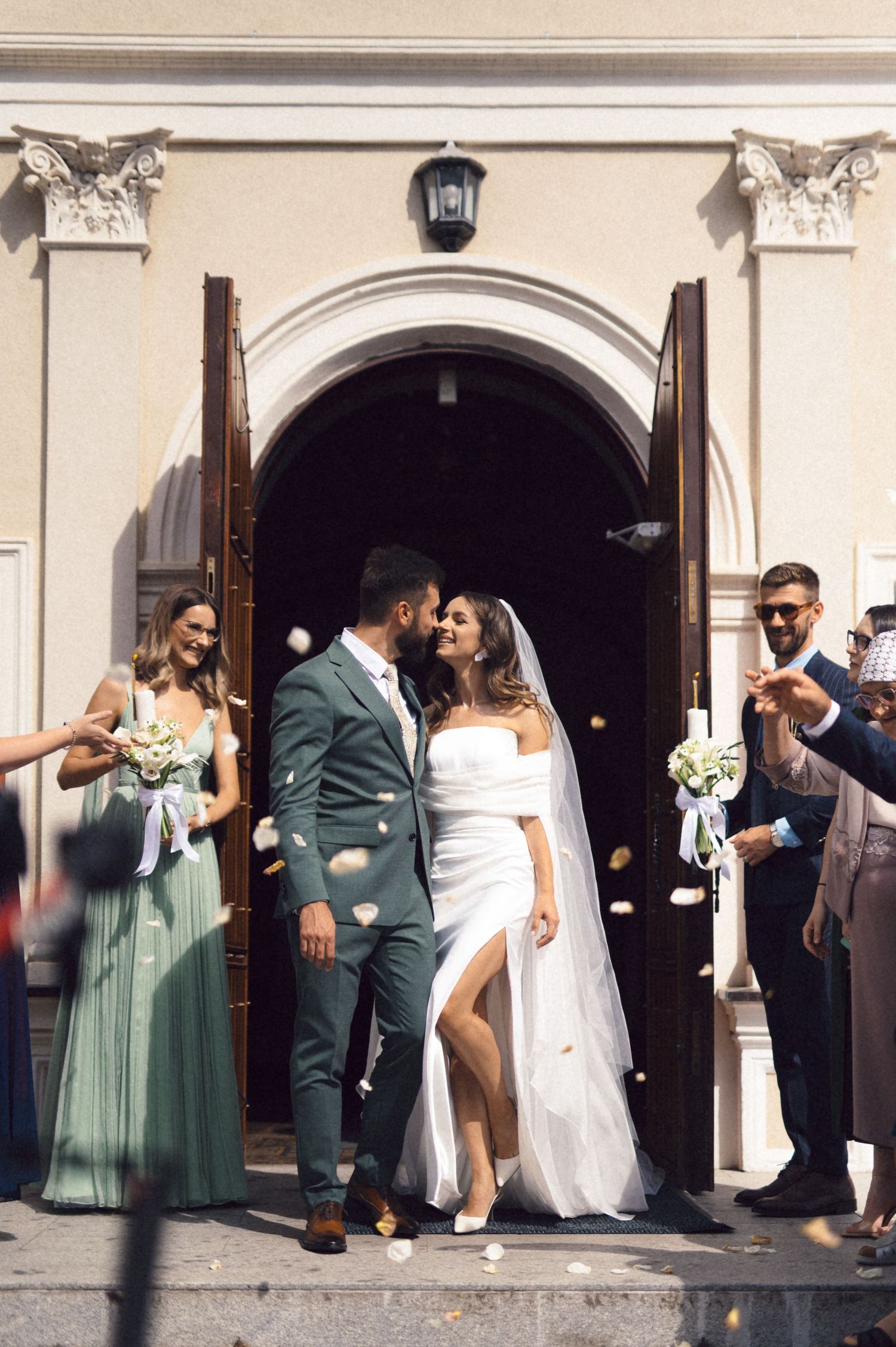 Wedding party members in sage green dresses and dark suits pose together in an elegant arched colonnade.