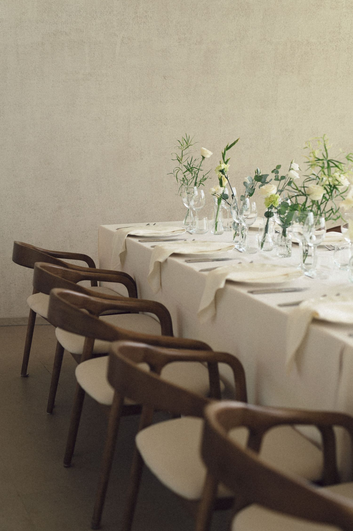 Elegant restaurant table settings with white linens and wooden chairs arranged in a row against a neutral wall.