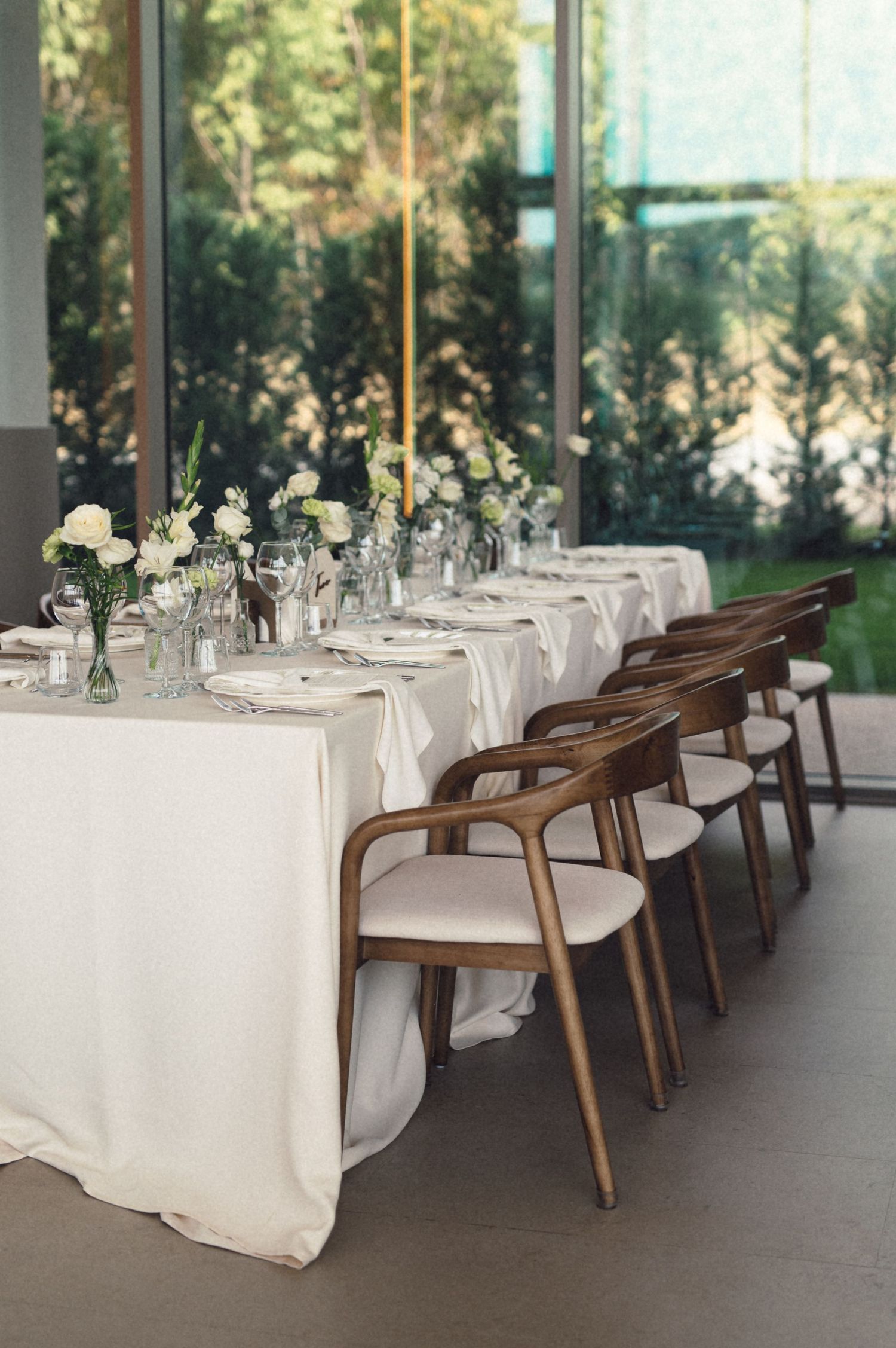 Elegant wedding reception tables with white linens, wooden chairs, and floral centerpieces arranged in a row by large windows.