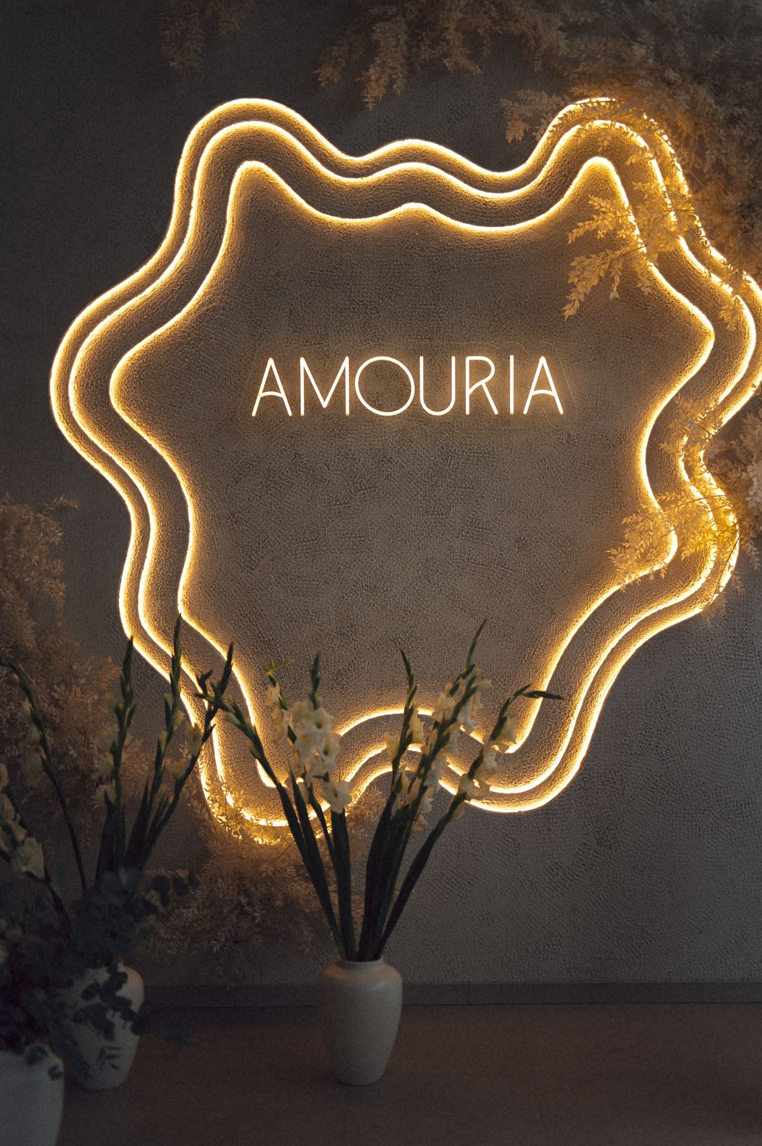 Illuminated neon light art installation spells out 'AMOURIA' against a dark wall with potted plants below.