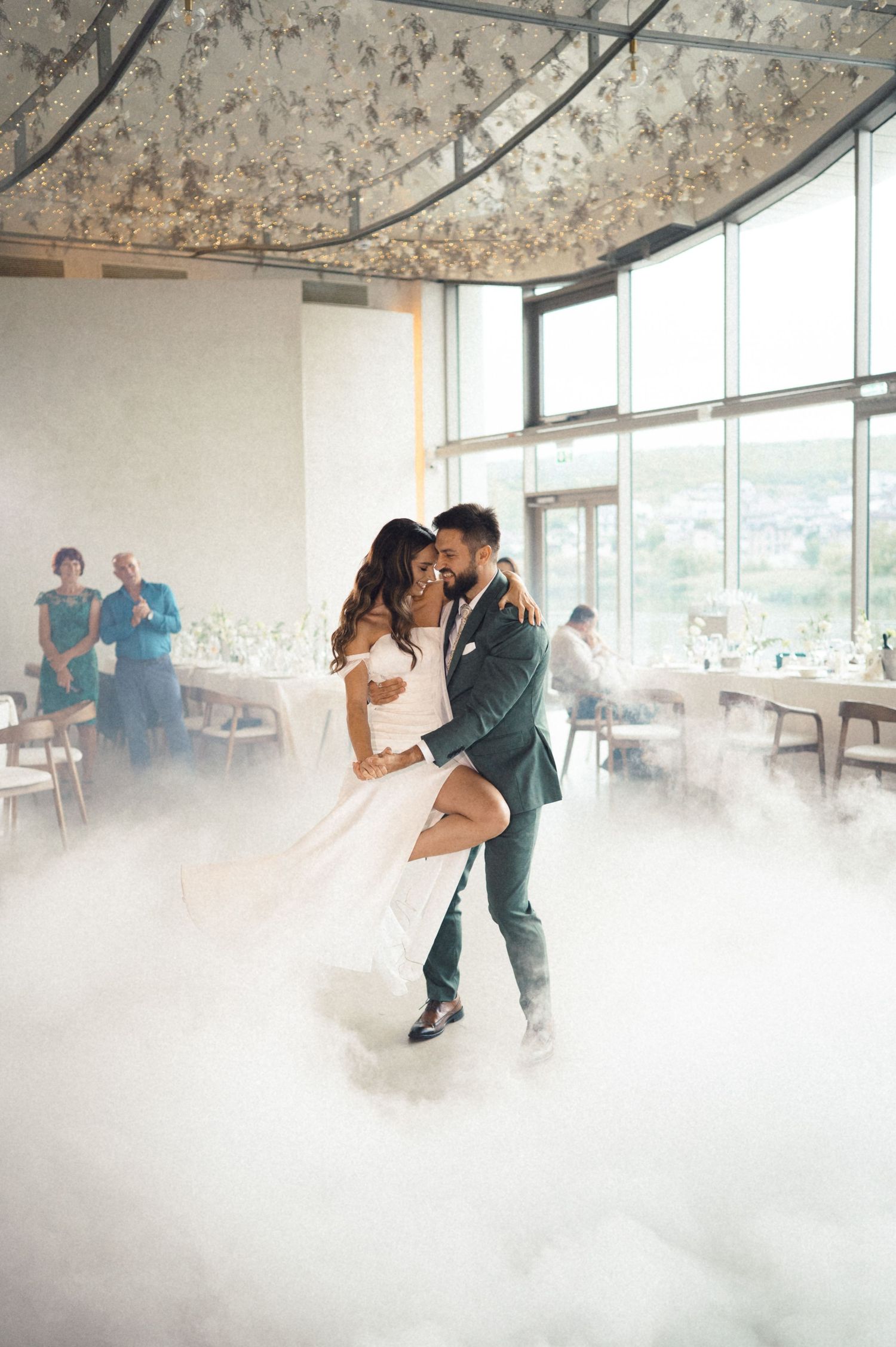 A couple dances together in an elegant room filled with atmospheric fog during their romantic first dance at a wedding.