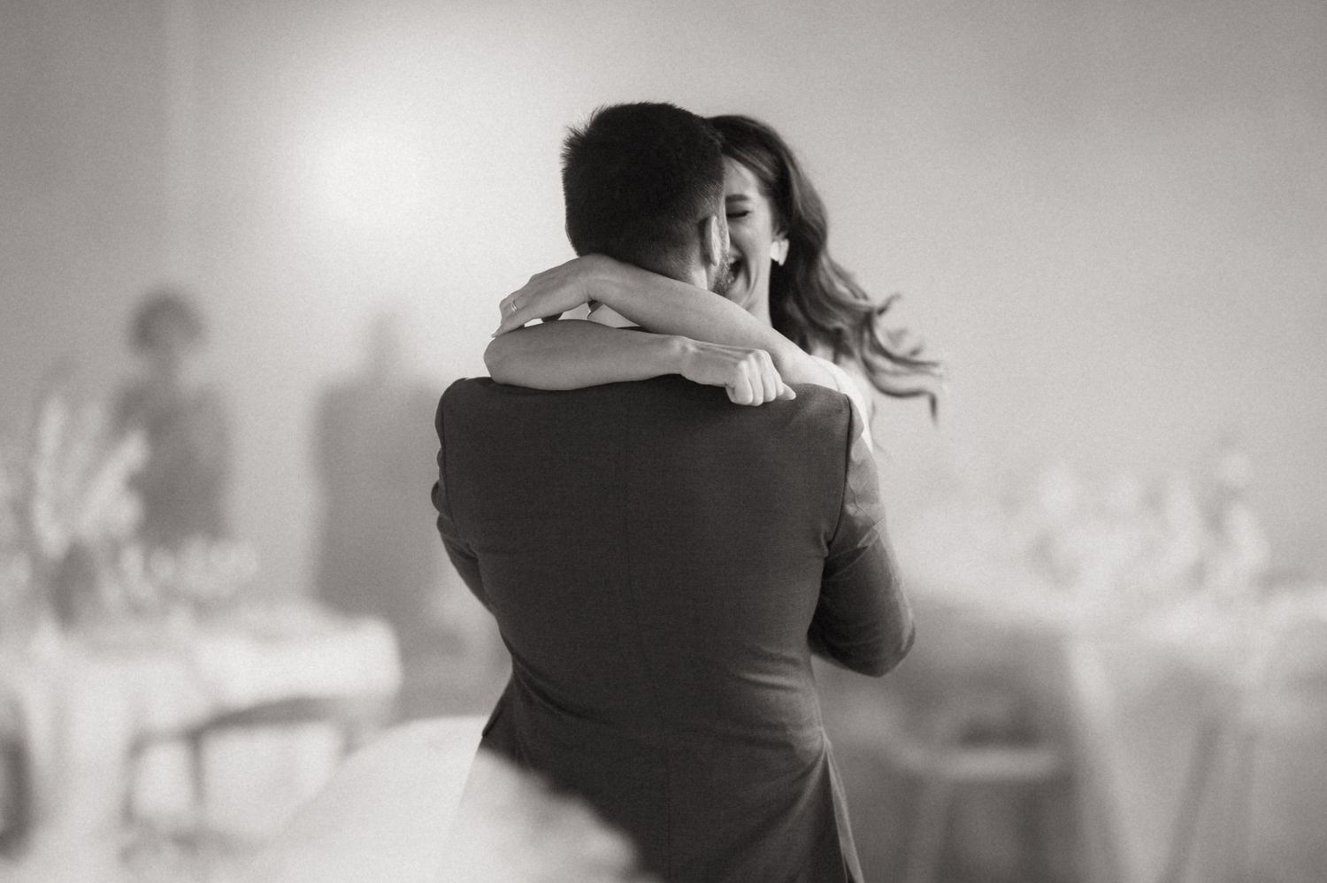 Black and white photo of a couple embracing and dancing at a wedding reception with other guests in the background.