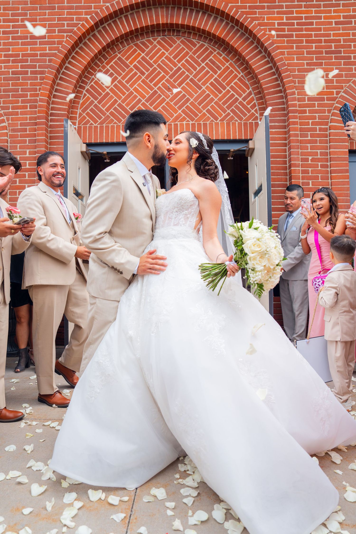 Wedding couple dips dramatically against brick archway as guests celebrate.