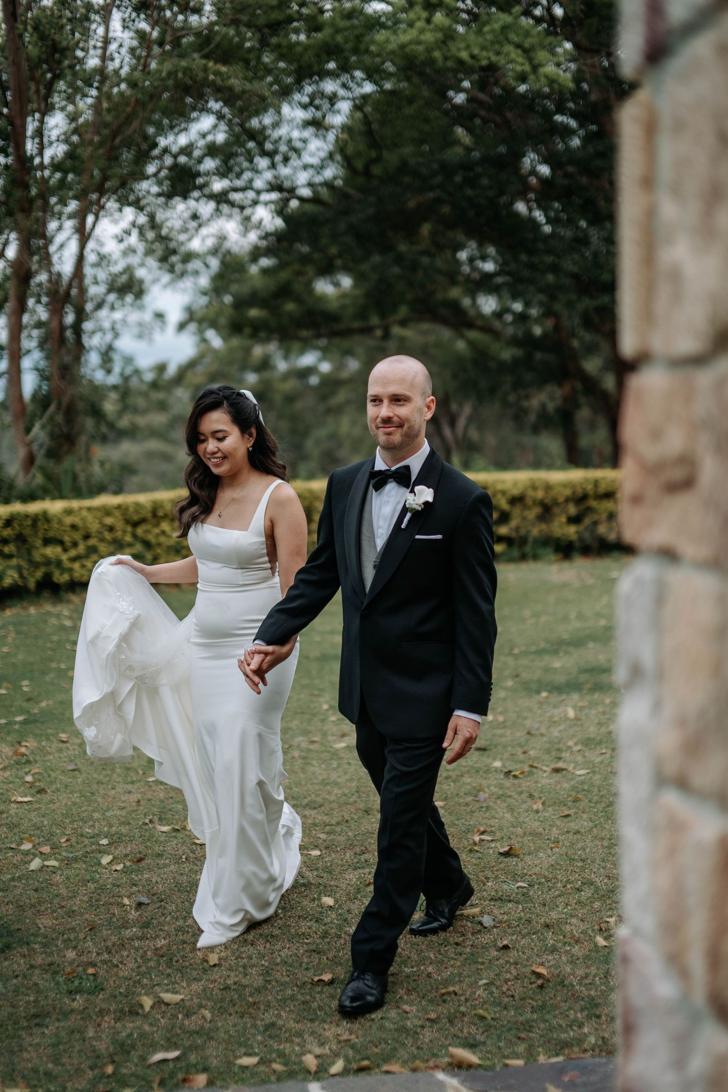 A bride and groom take playful dance steps together outdoors on their wedding day in a tree-lined setting.