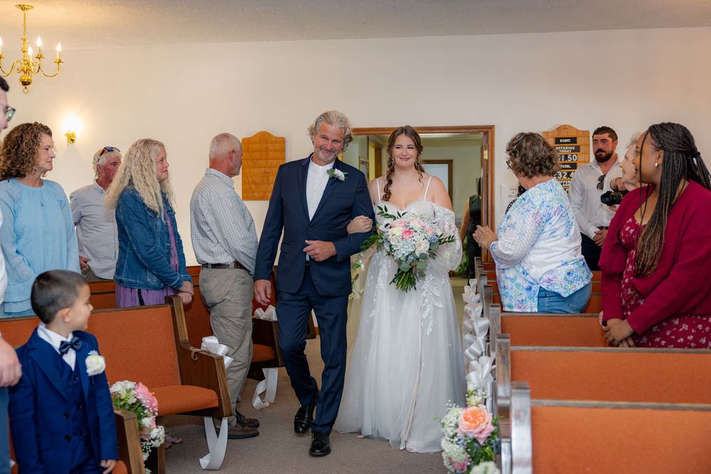 A small chapel wedding ceremony with guests seated in pews while a couple walks down the aisle decorated with flowers.